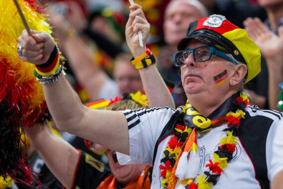 Ein leidenschaftlicher deutscher Fußballfan, der einen geschmückten Hut, eine Sonnenbrille und Deutschland-Accessoires trägt, jubelt in einer Menschenmenge, die mit der deutschen Flagge geschminkt und festlich gekleidet ist.