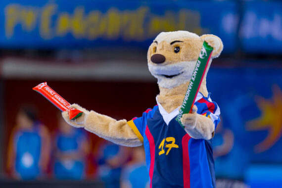 Ein kostümiertes Maskottchen in einem blauen Sporttrikot hält in jeder Hand ein rotes und ein grünes Banner und tritt vor einer verschwommenen Menschenmenge und einem blauen Hintergrund energisch auf.