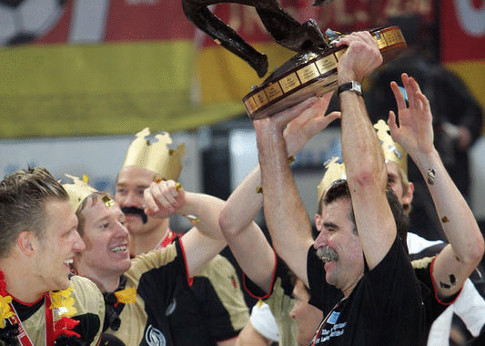 Eine Gruppe von Menschen mit goldenen Papierkronen feiert, während ein Mann mit Schnurrbart eine Trophäe über seinen Kopf hebt. Konfetti fällt um sie herum, und alle lächeln und jubeln.