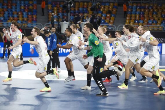 Eine Gruppe männlicher Handballspieler in weißen Trikots läuft freudig über ein Hallenfeld und jubelt mit einem Lächeln und erhobenen Armen. Die Zuschauer sitzen auf den bunten Tribünen im Hintergrund.