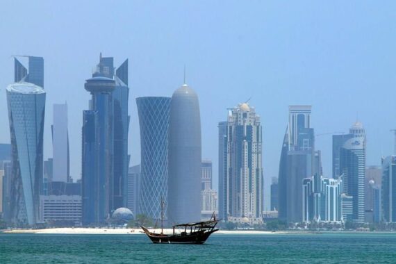 Ein traditionelles Holzboot schwimmt auf türkisfarbenem Wasser mit modernen Wolkenkratzern und Gebäuden der Skyline von Doha im Hintergrund unter einem klaren blauen Himmel.