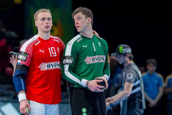 Zwei männliche Handballspieler stehen zusammen auf dem Spielfeld, einer in einem roten Trikot mit der Nummer 19, der andere in einem grünen Torwarttrikot. Der Spieler in Rot hat seinen Arm um die Schulter des Torhüters gelegt.