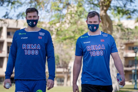 Zwei Männer in blauen Sportuniformen und schwarzen Gesichtsmasken gehen im Freien spazieren, jeder hält eine Wasserflasche. Im sonnigen Hintergrund sind Bäume und Gebäude zu sehen. Auf ihren Hemden steht REMA 1000 und sie tragen die norwegische Flagge.