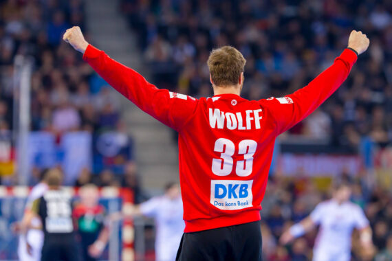 Ein Handballspieler, der ein rotes Trikot mit dem Namen Wolff und der Nummer 33 trägt, reißt beide Arme jubelnd in die Höhe, während im Hintergrund Spieler und Zuschauer zu sehen sind.