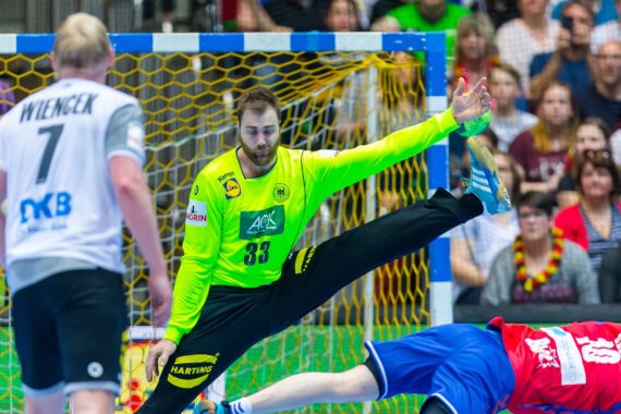 Ein Handballtorwart in einem leuchtend gelben Trikot streckt sich, um einen Schuss abzuwehren, während ein Spieler in Blau zu Boden fällt. Ein anderer Spieler in Weiß steht in der Nähe. Die Zuschauer im Hintergrund schauen aufmerksam zu.