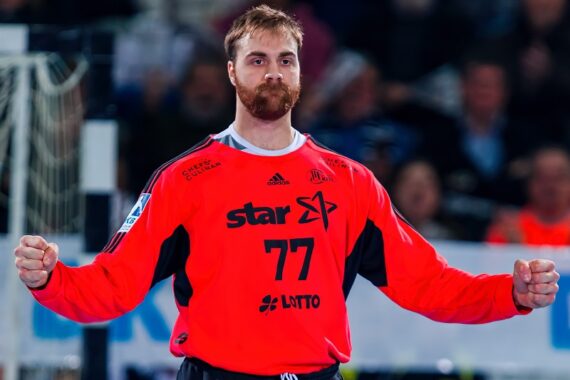 Ein Handballspieler mit Bart und Schnurrbart, der ein leuchtend rotes Trikot mit der Nummer 77 und Sponsorenlogos trägt, steht mit geballten Fäusten in einer Sportarena. Der Hintergrund ist unscharf mit Zuschauern.