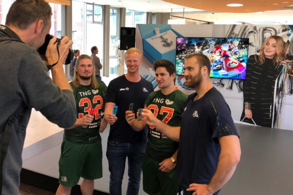 Vier Männer stehen zusammen und posieren für ein Foto, drei in grünen Sportuniformen und einer in Freizeitkleidung, in der Hand Getränke. Ein Fotograf macht ein Foto von ihnen. Hinter ihnen sind ein bunter Hintergrund und das Plakat einer lächelnden Frau zu sehen.