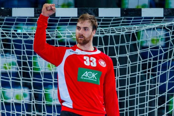 Ein männlicher Handballspieler in einem roten Trikot mit der Nummer 33 hebt vor einem Tornetz die Faust, um zu feiern oder ein Zeichen zu setzen.