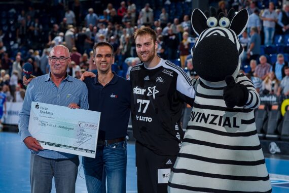 Vier Personen stehen auf einem Sportplatz: ein Mann mit einem großen Scheck, ein weiterer Mann, ein Sportler in einem schwarzen Trikot mit der Nummer 77 und eine Person in einem Zebra-Maskottchenkostüm. Im Hintergrund ist ein Publikum zu sehen.