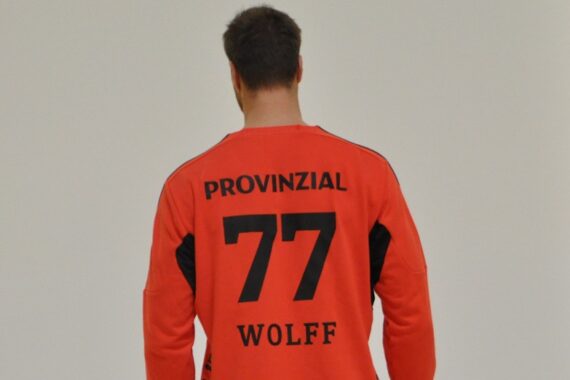 Eine Person, die ein rotes Sporttrikot mit dem Namen Wolff, der Nummer 77 und der Provinzial auf dem Rücken trägt, steht vor einem schlichten hellen Hintergrund.