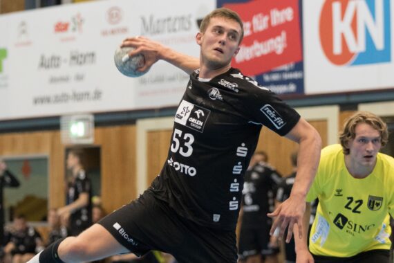 Ein männlicher Handballspieler in einem schwarzen Trikot mit der Nummer 53 ist mitten in der Aktion und bereitet sich auf den Wurf vor. Ein anderer Spieler in einem gelben Trikot ist im Hintergrund auf einem Hallenplatz zu sehen.