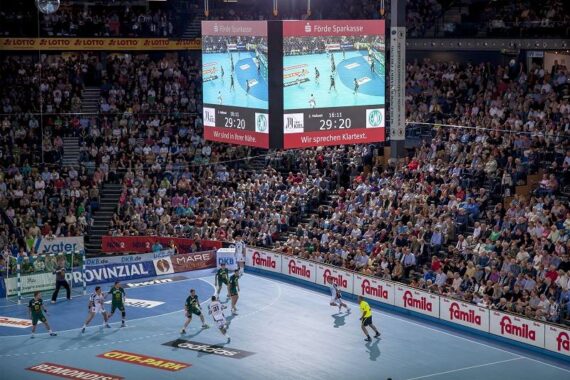 Eine volle Halle mit Zuschauern, die ein Handballspiel verfolgen. Zwei große Bildschirme zeigen den Spielstand an: 29:20. Spieler in grün-weißen Trikots sind auf dem blauen Spielfeld aktiv. Verschiedene Werbungen säumen die Arena.