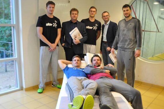 Sechs Männer posieren in einem fitnessstudioähnlichen Raum. Vier von ihnen stehen im hinteren Teil und halten Dokumente in der Hand, sie tragen Freizeit- oder Geschäftskleidung. Zwei Männer in Sportkleidung liegen auf einem medizinischen Untersuchungstisch im vorderen Bereich und wirken entspannt und lächelnd.