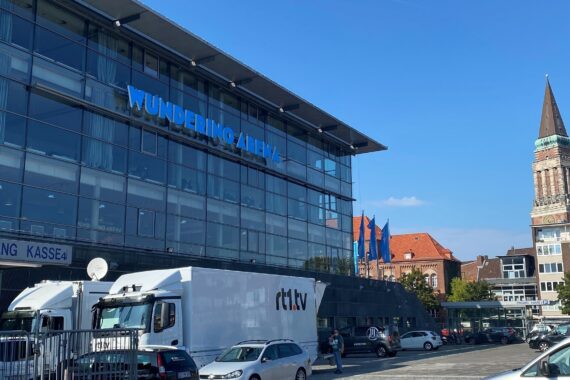 Zu sehen ist ein modernes Glasgebäude mit dem Schriftzug Wunderino Arena. Davor sind geparkte Fahrzeuge und ein NDR-Fernsehwagen zu sehen. Auf der rechten Seite erhebt sich ein hoher Backsteinturm in den strahlend blauen Himmel.