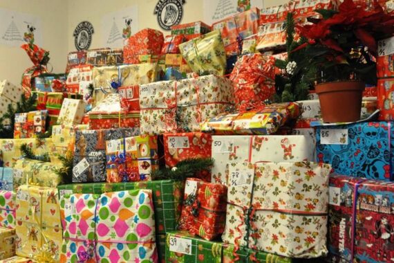Auf den Tischen stapeln sich bunte, verpackte Weihnachtsgeschenke mit verschiedenen Mustern, einige sind mit grünen Girlanden und einem roten Weihnachtsstern in einem Topf auf der rechten Seite geschmückt.