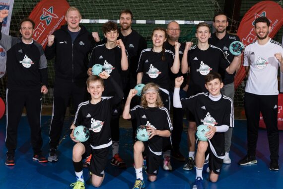 Eine Jugendhandballmannschaft posiert in einer Halle mit ihren Trainern, die passende schwarze Uniformen tragen und Handbälle in der Hand halten. Sie lächeln und recken die Fäuste zum Jubel. Im Hintergrund sind Banner und ein Netz zu sehen.