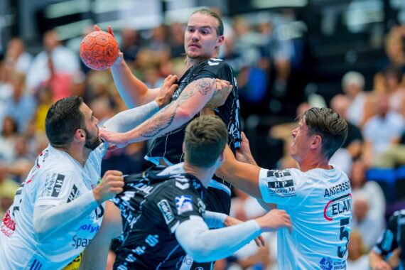 Ein Handballspieler in einem schwarzen Trikot springt, um den Ball zu werfen, während drei Spieler in weißen Trikots versuchen, ihn zu blockieren. Die überfüllte Arena im Hintergrund ist leicht unscharf.