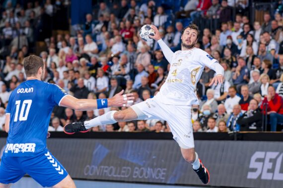 Ein Handballspieler in einem weißen Trikot springt in die Luft, um den Ball zu werfen, während ein Spieler in einem blauen Trikot mit der Nummer 10 versucht, ihn zu blocken. Eine große, konzentrierte Menge schaut von der Tribüne aus zu.