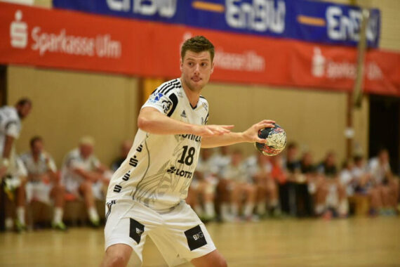 Ein männlicher Handballspieler in einem weißen Trikot bereitet sich darauf vor, den Ball während eines Spiels zu passen, während seine Mannschaftskameraden im Hintergrund sitzen und Sponsorenbanner an der Wand zu sehen sind.