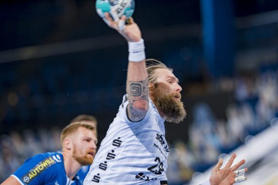 Ein bärtiger Handballspieler in einem weißen Trikot bereitet sich auf den Wurf vor, während ein Spieler in einem blauen Trikot während eines Spiels in einer Halle dicht hinter ihm verteidigt.