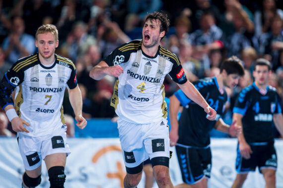 Zwei männliche Handballspieler in weißen Trikots feiern auf dem Spielfeld, einer schreit mit ausgestreckten Armen. Im Hintergrund sind Mannschaftskameraden und Gegner in blauen Trikots zu sehen, dahinter eine jubelnde Menge.