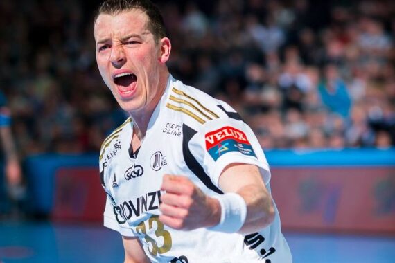 Ein männlicher Handballspieler in einem weißen Trikot reagiert während eines Spiels in einer mit Zuschauern gefüllten Halle leidenschaftlich, ballt die Faust und schreit.