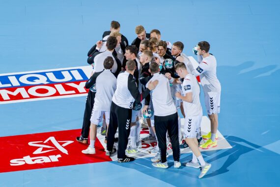 Eine Herren-Handballmannschaft in weißen und schwarzen Trikots kauert auf einem blauen Hallenplatz neben Logos mit der Aufschrift Liqui Moly und Star.