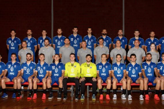 Eine Handballmannschaft in blauen Trikots und der Trainerstab stellen sich in einer Halle für ein Gruppenfoto auf. Drei Spieler in gelben Torwarttrikots sitzen in der ersten Reihe, weitere Spieler und Betreuer sind in Reihen hinter ihnen angeordnet.