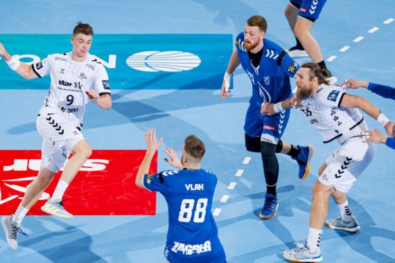 Ein Handballspiel in Aktion: Ein Spieler in einem weißen Trikot springt, um den Ball zu werfen, während seine Gegner in blauen Trikots den Ball verteidigen, während seine Mannschaftskameraden und andere Verteidiger auf einem blauen Hallenplatz zusehen.