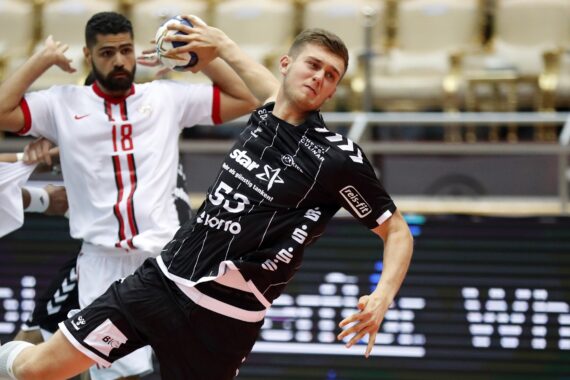 Ein Handballspieler in einem schwarzen Trikot ist im Begriff, den Ball zu werfen, während er während eines Spiels von einem Gegner in einem weiß-roten Trikot eng verteidigt wird. Der Hintergrund zeigt leere Sitzplätze in der Arena.
