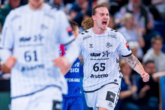 Ein männlicher Handballspieler in einem weißen Trikot mit der Nummer 65 jubelt mit offenem Mund und geballter Faust auf dem Spielfeld. Ein weiterer Spieler in Weiß und einer in Blau sind im Hintergrund zu sehen. Die Zuschauer auf der Tribüne schauen zu.