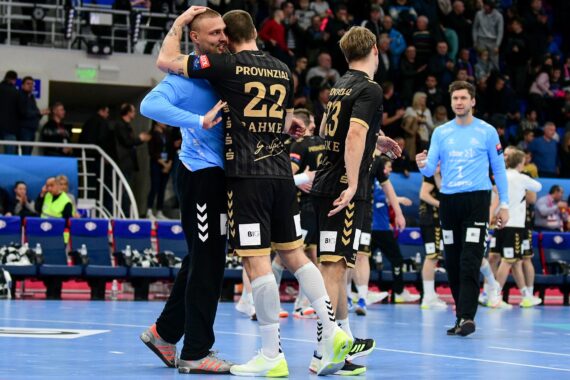 Zwei Handballspieler in schwarzen Trikots umarmen sich auf einem Hallenplatz und feiern. Andere Mannschaftskameraden und Offizielle sind zu sehen; Zuschauer schauen von der Tribüne im Hintergrund zu. Die Szene vermittelt den Eindruck eines siegreichen oder emotionalen Moments.