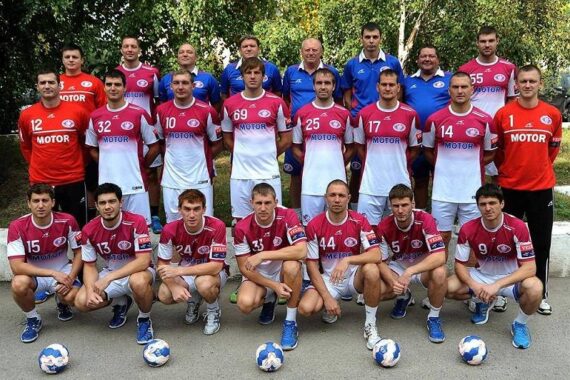 Eine Herren-Handballmannschaft stellt sich im Freien in drei Reihen auf und trägt passende rot-weiß-rosa Trikots mit Motor auf der Vorderseite. Vier Trainer in Rot oder Blau stehen im Hintergrund. Mehrere Handbälle liegen auf dem Boden. Im Hintergrund sind Bäume zu sehen.