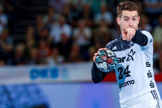 Ein männlicher Handballspieler in einem weiß-schwarzen Trikot mit der Nummer 24 hält während eines Spiels einen Handball in der rechten Hand und zeigt mit der linken Hand nach vorne, während im Hintergrund eine verschwommene Menschenmenge zu sehen ist.