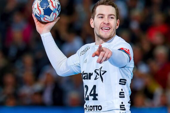 Ein männlicher Handballspieler, der ein weißes Trikot mit der Nummer 24 trägt, hält einen Handball in der rechten Hand und zeigt mit der linken Hand nach vorne, während er lächelt. Im Hintergrund ist eine unscharfe Menschenmenge zu sehen.