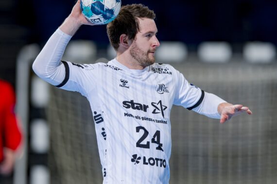 Ein männlicher Handballspieler in einem weißen Trikot mit der Nummer 24 bereitet sich darauf vor, einen blau-weißen Ball während eines Spiels zu werfen. Der Hintergrund ist unscharf, ein Tornetz und ein weiterer Spieler in Rot sind zu sehen.