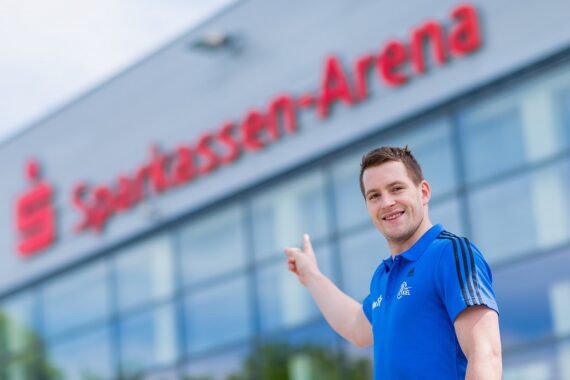 Ein lächelnder Mann in einem blauen Hemd zeigt auf ein großes Gebäude, auf dessen Fassade in roten Buchstaben Sparkassen-Arena steht. Das Gebäude hat reflektierende Glasfenster.
