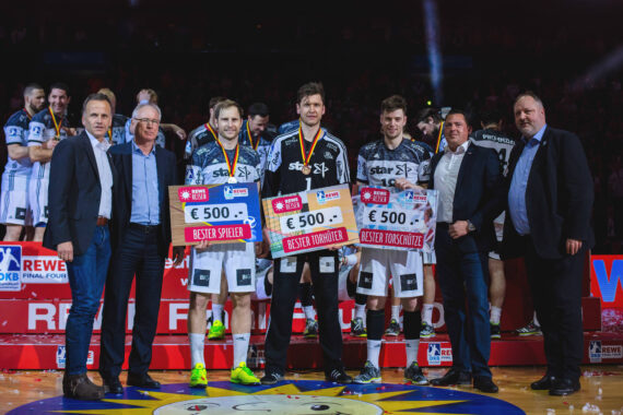 Drei Handballer in schwarz-weißen Uniformen halten große Schecks und Medaillen in der Hand und stehen mit sechs formell gekleideten Männern auf einer Bühne mit einer Menschenmenge und heller Beleuchtung im Hintergrund.