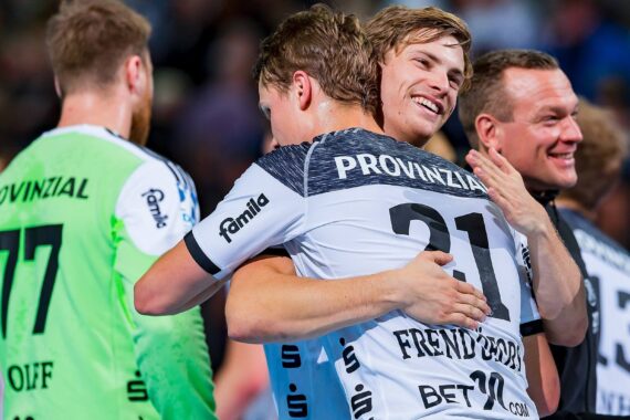Handballspieler in schwarz-weißen Trikots feiern auf dem Spielfeld, umarmen sich und lächeln. Auf dem Trikot eines Spielers steht die Nummer 21 und der Name Provinzial. Der Hintergrund ist unscharf und es sind mehrere Personen zu sehen.