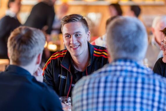 Ein junger Mann in einer Sportjacke lächelt und unterhält sich mit zwei anderen Personen an einem Tisch in einem belebten Innenbereich. Die Szene wirkt freundlich und gesellig.