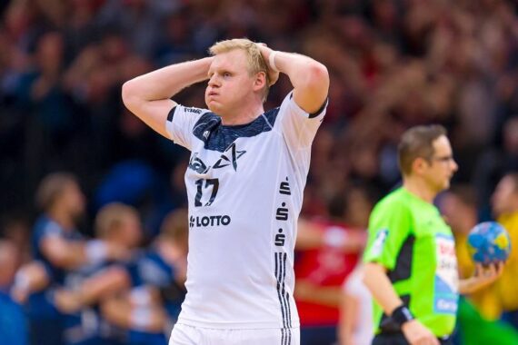 Ein männlicher Handballspieler in einem weißen Trikot mit der Nummer 17 steht mit den Händen auf dem Kopf und sieht frustriert aus. Ein Offizieller in einem grünen Hemd hält einen Ball, und im Hintergrund ist eine unscharfe Menschenmenge zu sehen.