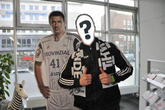 Eine Person mit einem Papier mit einem großen Fragezeichen im Gesicht steht in einem Raum und zeigt zwei Daumen nach oben neben einer lebensgroßen Pappfigur eines Sportspielers in einem weißen Trikot. In der Nähe befindet sich eine kleine Zebra-Statue.