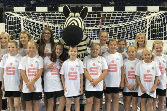 Eine Gruppe junger Mädchen in weißen Hemden steht vor einem Handballtor mit einer Person in einem Zebra-Maskottchen-Kostüm und lächelt für ein Gruppenfoto in einer Halle.