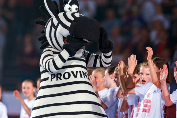 Eine Person in einem gestreiften Zebra-Maskottchenkostüm mit der Aufschrift PROVINZ steht inmitten einer Gruppe lächelnder Kinder in weißen Hemden; die Kinder klatschen und jubeln bei einer Indoor-Veranstaltung.