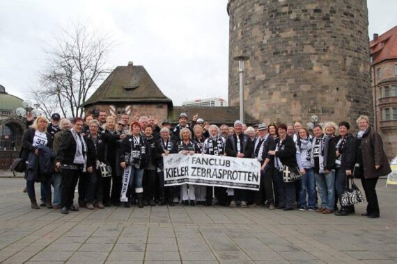 Eine große Gruppe von Menschen steht im Freien zusammen und hält ein Transparent mit der Aufschrift Fanclub Kieler Zebrasprotten. Viele tragen schwarze und weiße Schals und Kleidung, im Hintergrund ist ein Steinturm zu sehen.