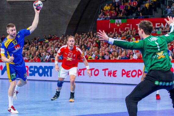 Ein Handballspieler in einem blau-gelben Trikot bereitet sich auf den Wurf vor, während ein Torwart in Grün und ein gegnerischer Spieler in Rot den Ball verteidigen, während die Zuschauer in der Arena zusehen.