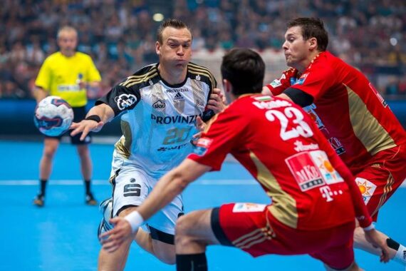 Ein Handballspieler in einem weißen Trikot bereitet sich auf den Wurf vor, während zwei Spieler in Rot ihn während eines Spiels abwehren.