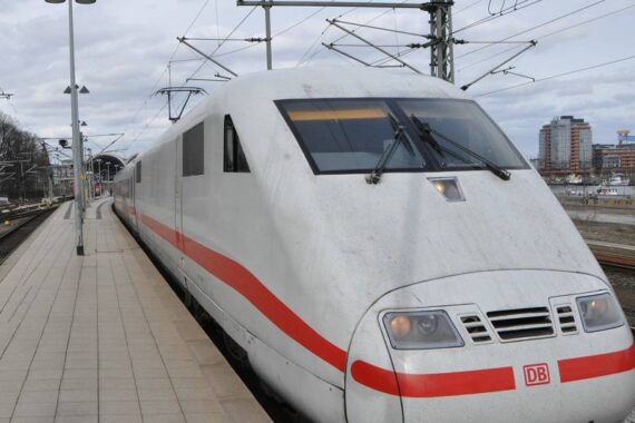 Ein weißer Hochgeschwindigkeitszug der Deutschen Bahn (DB) mit rotem Streifen hält an einem Bahnsteig in einem modernen Bahnhof im Freien. Im Hintergrund sind Gebäude und Oberleitungen zu sehen.
