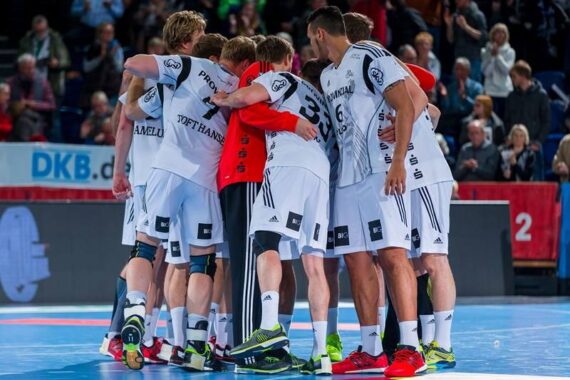 Eine Gruppe männlicher Handballspieler in weißen Trikots drängt sich auf einem Hallenplatz zusammen, mit Zuschauern im Hintergrund. Die Spieler haben ihre Arme umeinander gelegt, um die Einigkeit der Mannschaft vor einem Spiel zu demonstrieren.