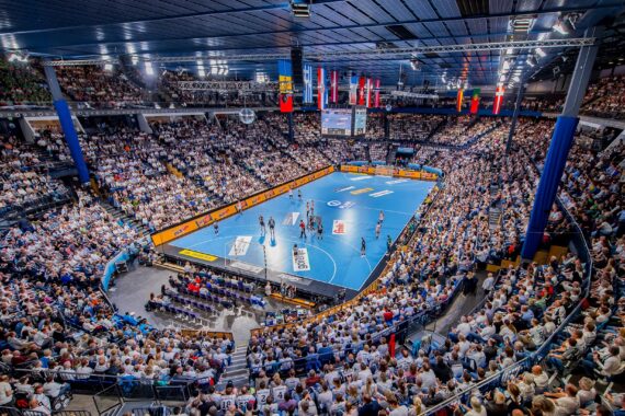 Eine überfüllte Halle mit Tausenden von Zuschauern, die ein Handballspiel auf einem hellblauen Spielfeld verfolgen, umgeben von Nationalflaggen und großen Videoleinwänden über dem Geschehen.